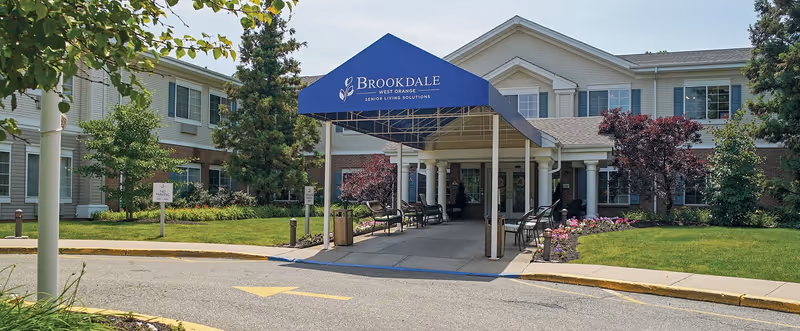 Entrance to Brookdale West Orange senior living facility with a blue canopy displaying the facility name. The building is two stories with white siding and brick accents, surrounded by landscaped greenery and trees.