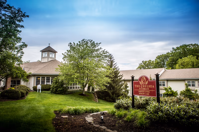 Exterior view of a senior living facility named The Chelsea at Forsgate, showing a large building with a cupola, surrounded by green trees and well-maintained landscaping under a partly cloudy sky.