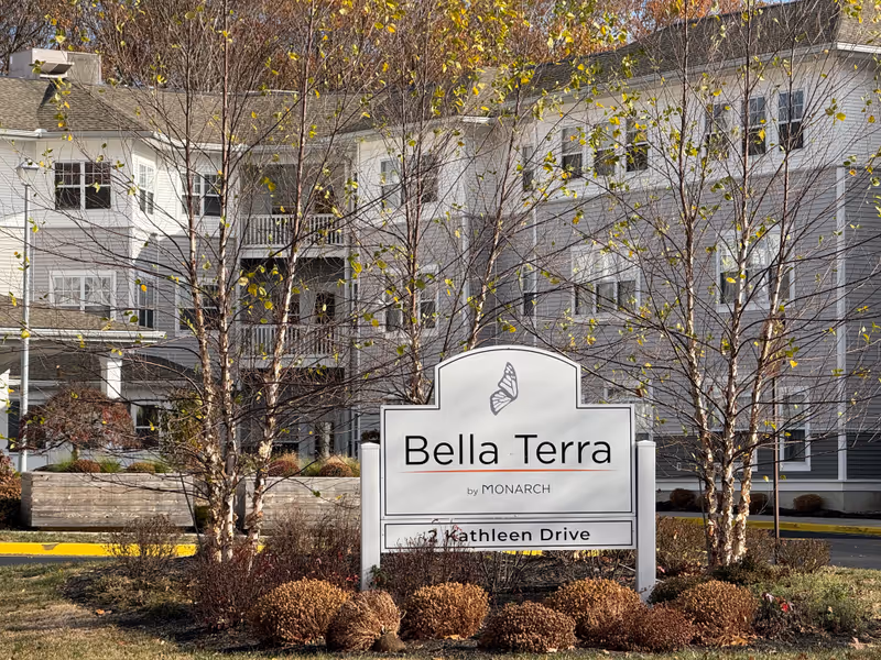 Outdoor view of the Monarch Bella Terra senior living facility sign with a butterfly logo, surrounded by trees and shrubs, with the building visible in the background.
