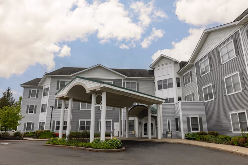 Front entrance of a multi-story gray senior living building with a covered porte-cochere and paved driveway.