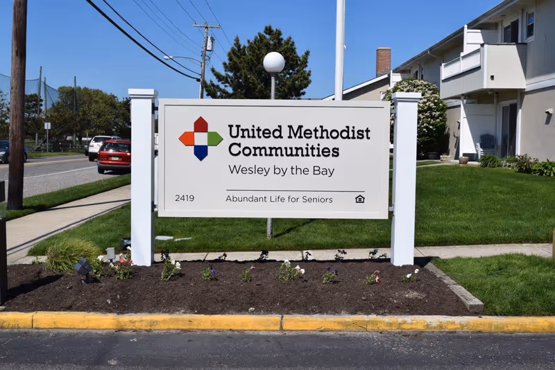 Outdoor view of a white sign for United Methodist Communities Wesley by the Bay, located on a landscaped area with small flowers and grass. The sign includes the tagline 'Abundant Life for Seniors' and the number 2419. In the background, there is a sidewalk, a road with cars, utility poles, and part of a building with balconies.