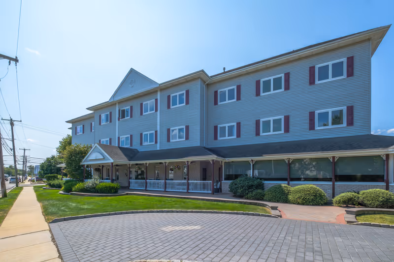 Exterior view of a three-story senior living facility building with gray siding and maroon shutters. The building has a covered porch with white railings and hanging flower baskets. There is a paved driveway with an arrow pointing forward and a well-maintained lawn with bushes and a sidewalk along the street.
