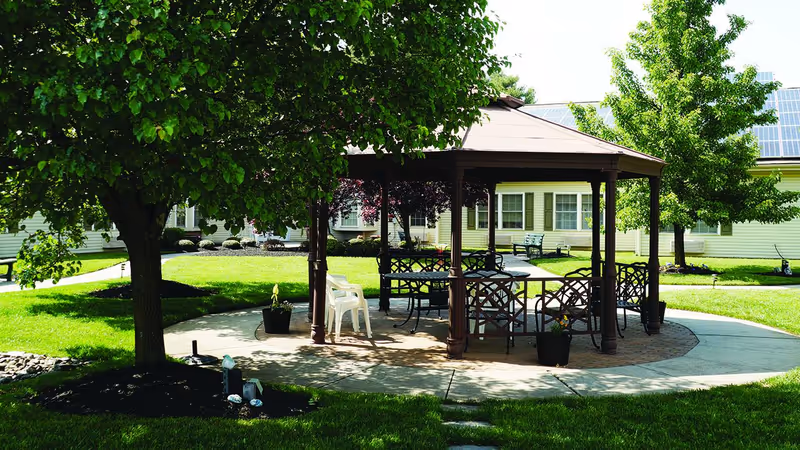 Outdoor gazebo with metal chairs and tables on a circular paved area surrounded by green grass, trees, and a building with solar panels on the roof in the background.