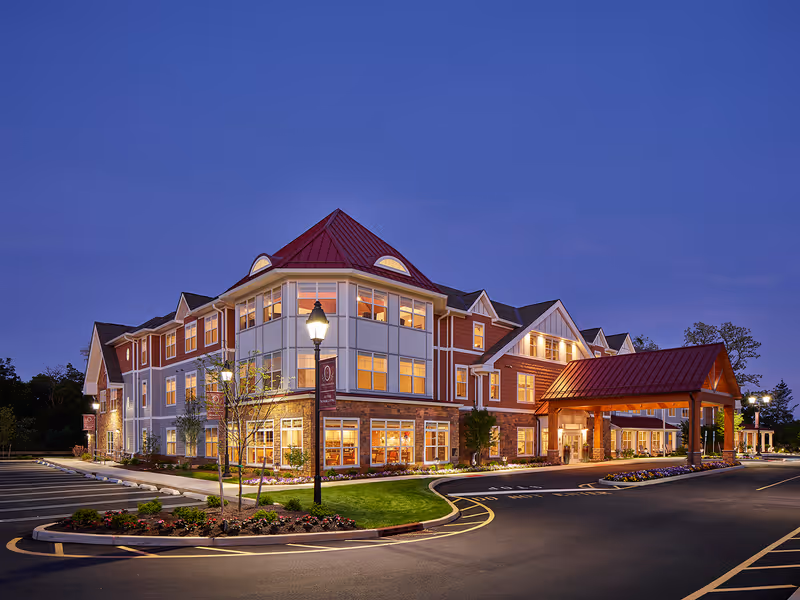 Exterior view of The Chelsea at Shrewsbury senior living facility at dusk, showing a large multi-story building with a red roof, well-lit windows, a covered entrance, landscaped flower beds, and a parking area.