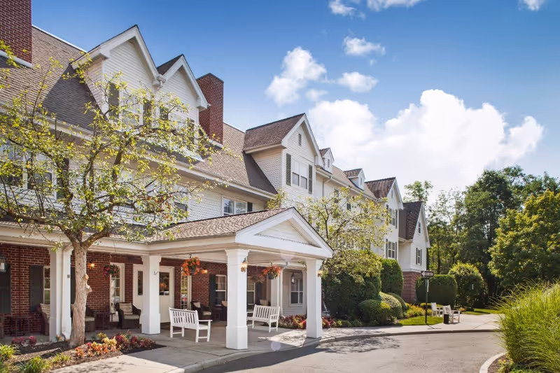 Exterior view of a senior living facility building with white siding and brick accents, featuring a covered entrance with white columns, benches, trees, and well-maintained landscaping under a partly cloudy blue sky.
