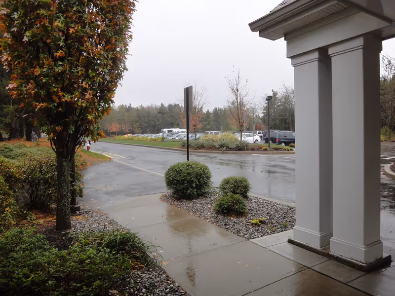 View from under a covered entrance looking out at a wet driveway, landscaped shrubs and a parking lot on a rainy day.