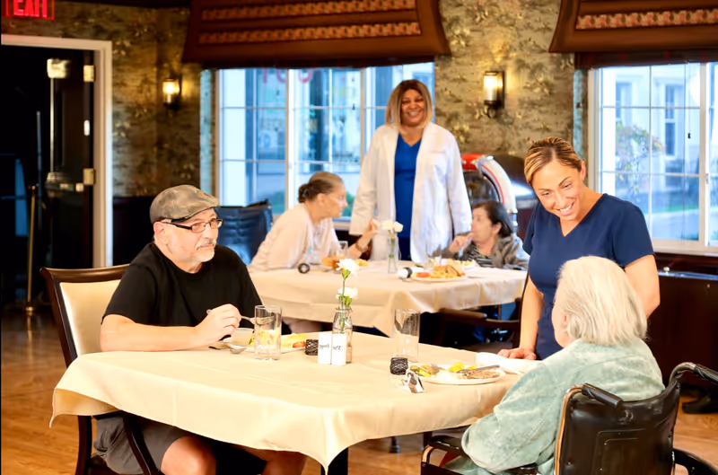 A dining room in a senior living facility where an elderly man is seated at a table eating, while a caregiver in navy scrubs smiles and interacts with an elderly woman in a wheelchair. In the background, another caregiver in a white coat stands near a table where two elderly women are seated and eating. The room has large windows, warm lighting, and a cozy atmosphere.