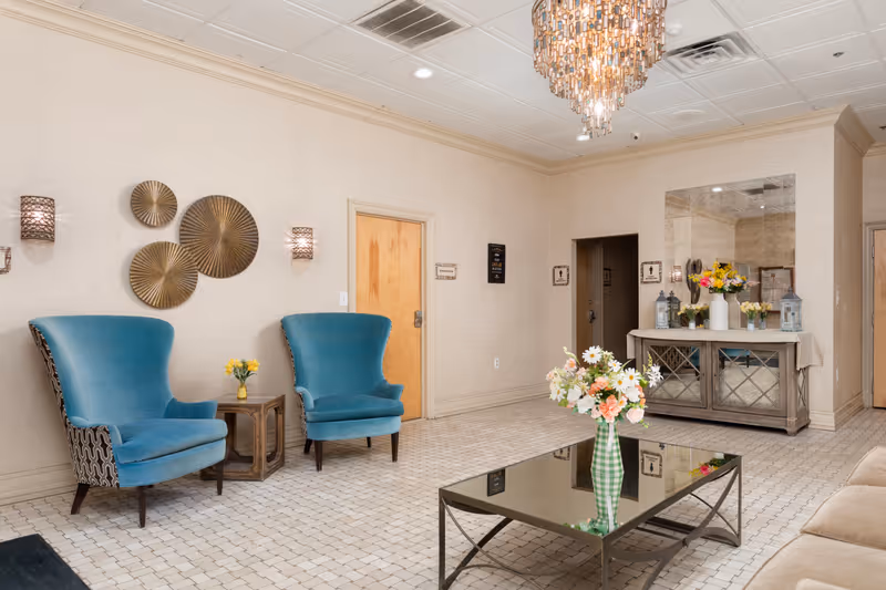 A bright and elegant common area with two blue upholstered armchairs, a small wooden side table with a yellow flower vase, a mirrored console table decorated with various flower arrangements and lanterns, a glass coffee table with a green checkered vase holding a bouquet of flowers, beige tiled floor, and a decorative chandelier hanging from the ceiling.