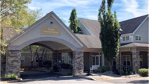 Exterior view of Stein Assisted Living facility showing the entrance with a covered driveway supported by stone pillars, surrounded by trees and shrubs under a clear sky.