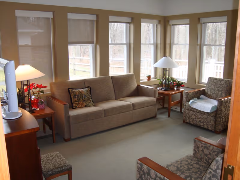 Sunny living room with a beige sofa, patterned armchairs, side tables, lamps, and several large windows.
