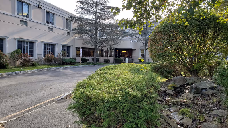 Exterior view of a senior living facility building with a driveway leading to the entrance. The building has multiple windows and is surrounded by landscaped greenery including bushes, trees, and rocks.