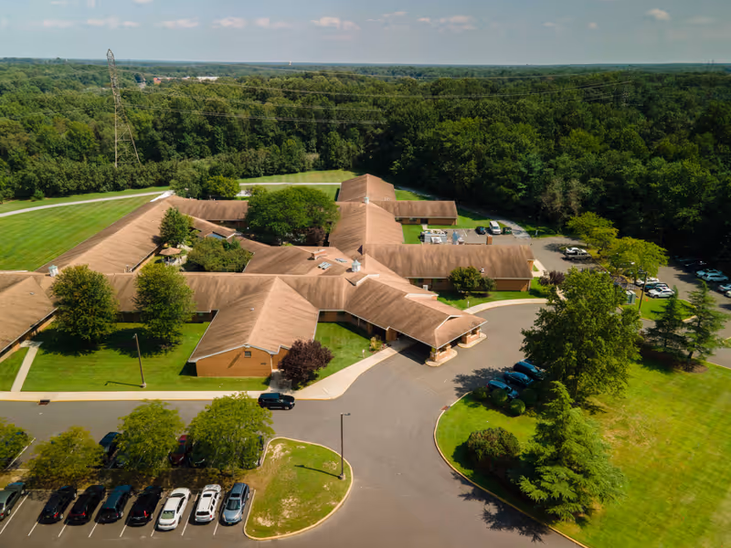 Aerial view of a single-story senior living facility with brown roofs surrounded by green lawns, trees, and a parking lot with several cars. The building is set against a backdrop of dense forest under a partly cloudy sky.