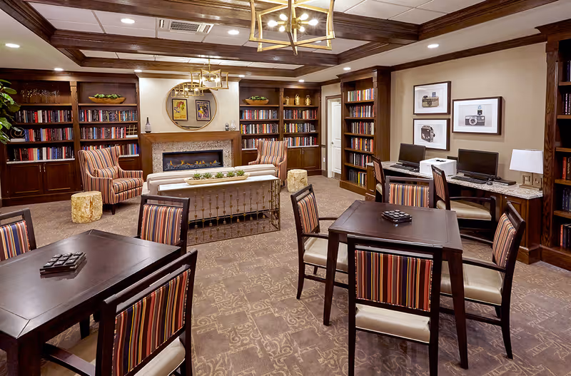 A cozy and well-lit common area featuring dark wood bookshelves filled with books, a modern fireplace with a round mirror and framed pictures above it, striped armchairs, and tables with striped chairs. There are also two computer workstations with chairs and framed camera pictures on the wall. The room has a patterned carpet and warm lighting from ceiling fixtures.
