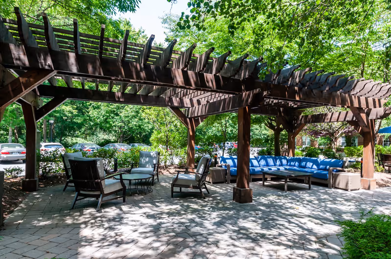 Outdoor seating area under a wooden pergola with cushioned chairs and a long blue cushioned sofa surrounded by trees and greenery, with a parking lot visible in the background.