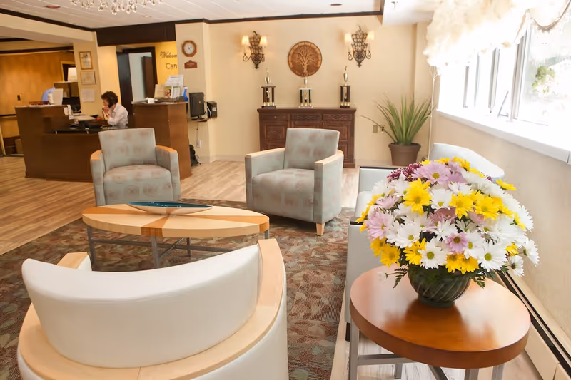 A bright senior living facility lounge with armchairs, a reception desk in the background, and a side table holding a vase of yellow and white flowers.