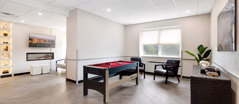 Bright communal lounge with a small pool table, chairs, a wall-mounted TV above a linear fireplace, and decorative shelving.