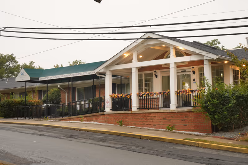 Exterior view of Complete Care at Chestnut Hill nursing and rehabilitation center showing a brick building with a covered entrance, white columns, and a ramp leading to the door. The entrance is decorated with fall-themed garlands and there are bushes and trees around the building.
