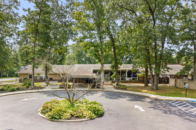 Front exterior view of a single-story building surrounded by tall trees and greenery. There is a circular driveway with a landscaped island in the center featuring a small tree and plants. The building has large windows and a covered entrance area.