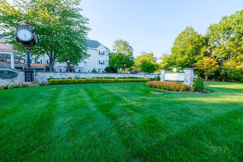 A well-maintained green lawn with a stone wall and flower beds in front of a senior living community building. There is a large decorative clock on a post to the left and a sign on the right that reads 'Brandywine Living at Summit A Senior Living Community'. Trees and a clear blue sky are visible in the background.