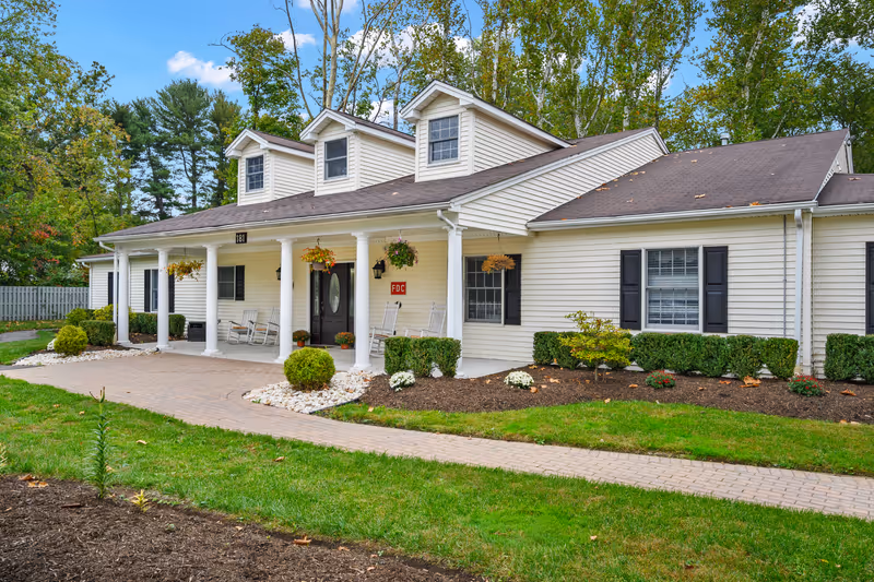 Front exterior of a single-story beige memory care building with a covered porch, white columns, rocking chairs, and landscaped grounds.