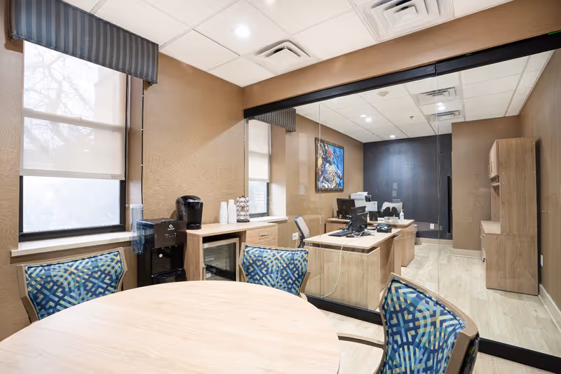 A small office space with a round wooden table and four patterned chairs in the foreground. Behind a glass partition is a desk with computer monitors, office chairs, and a printer. The room has beige walls, a window with a roller shade, a coffee maker, and a small refrigerator. A colorful framed artwork hangs on the wall behind the desk.