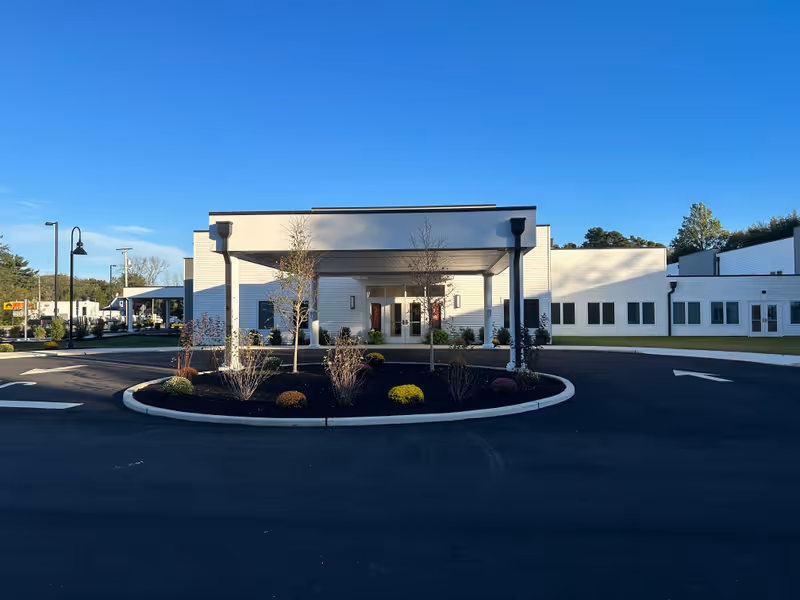 Front exterior view of New Standard Senior Living at Hammonton building with a covered entrance, landscaped roundabout with small trees and shrubs, clear blue sky, and paved driveway.