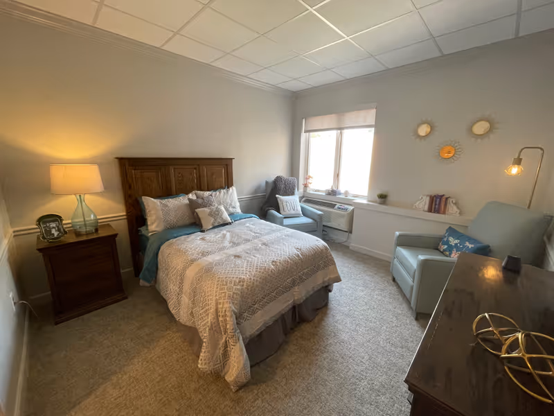 A cozy bedroom in a senior living facility featuring a wooden bed with patterned bedding, a nightstand with a lamp and picture frame, two armchairs near a window with a cushion that says 'LOVE', a dresser with decorative items, and three small round mirrors on the wall.
