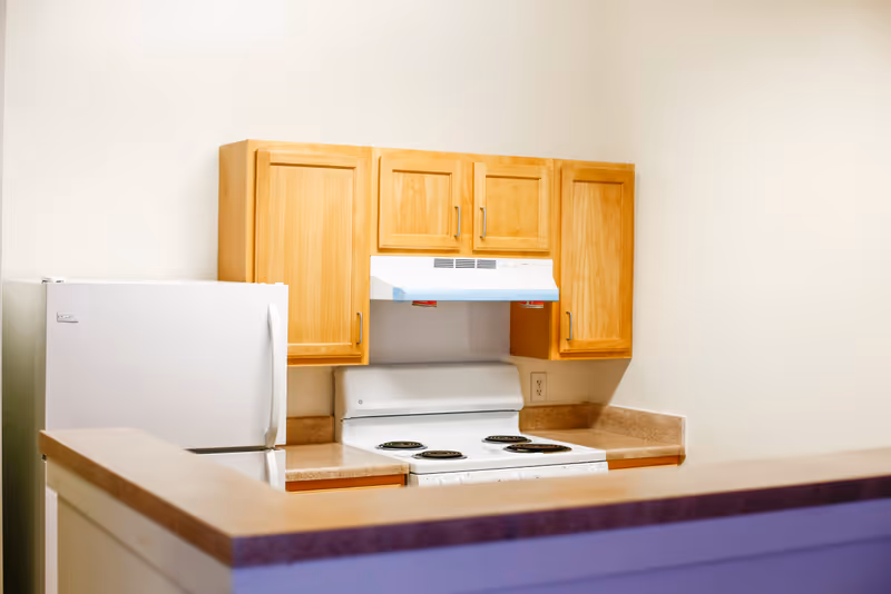 A small kitchen area with light wood cabinets, a white refrigerator, a white electric stove with four burners, and a white range hood. The countertop is beige, and the walls are painted white.