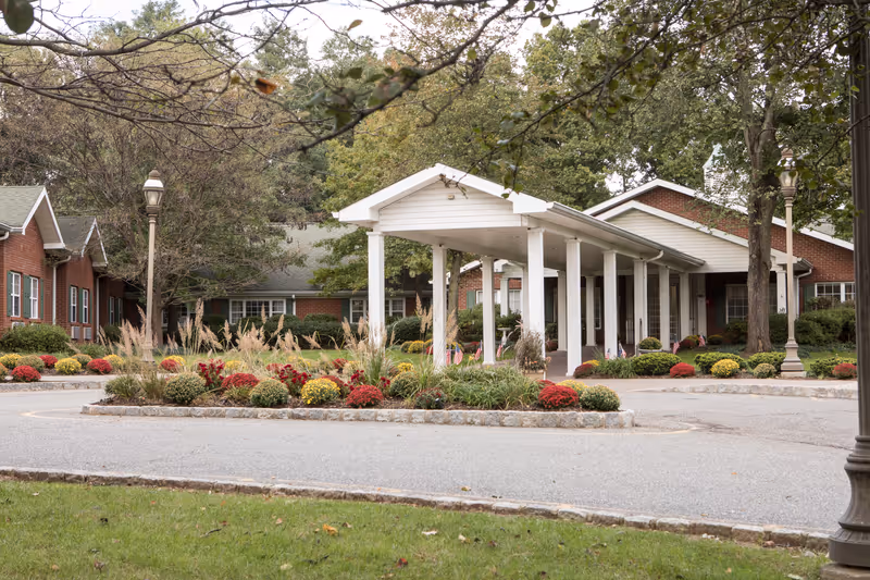 Entrance area of a senior living facility with a covered drop-off zone supported by white columns. The building is made of red brick with white trim and surrounded by landscaped gardens featuring bushes, flowers, and trees. There are street lamps and a circular driveway in front of the entrance.