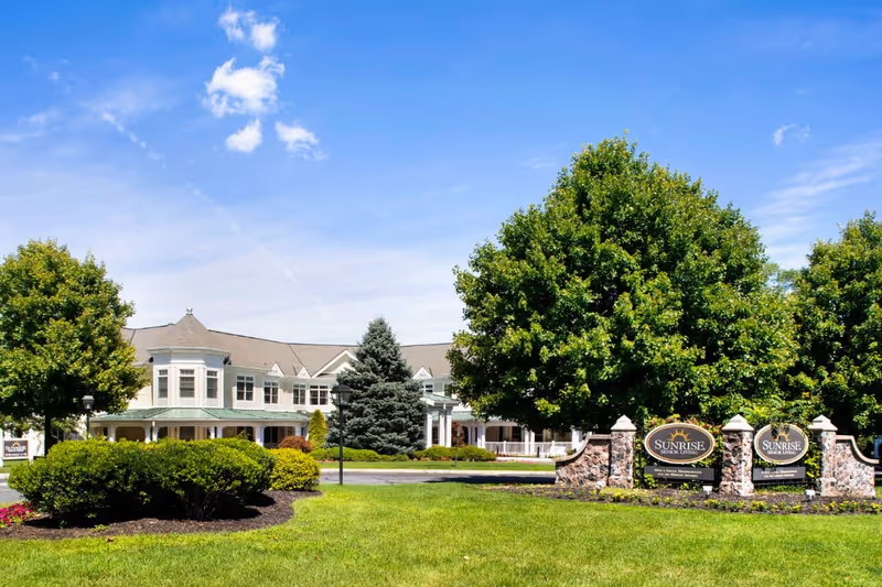 Exterior view of Sunrise of Madison senior living facility on a sunny day, showing a large building with white siding, multiple windows, and a green roof. The foreground features well-maintained green grass, bushes, trees, and a stone sign with the Sunrise Senior Living logo.