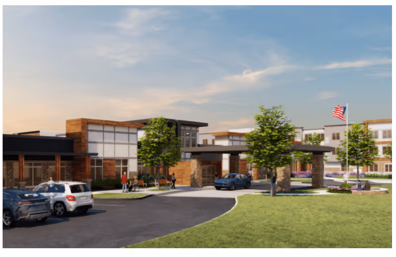 Exterior view of a modern senior living facility with a driveway, parked cars, trees, and an American flag on a flagpole. People are walking and sitting near the entrance and landscaped areas.