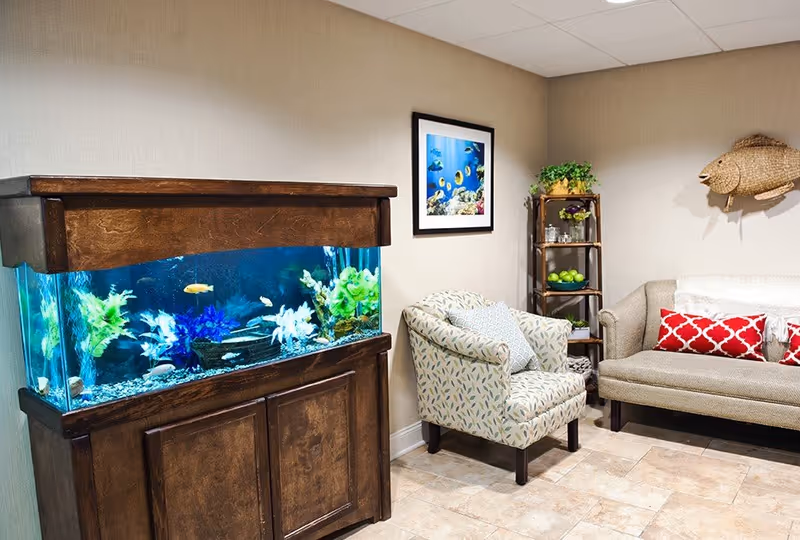 A cozy sitting area in a senior living facility featuring a large wooden aquarium filled with colorful fish, a patterned armchair with a cushion, a beige sofa with red and white pillows, a small wooden shelf with decorative items and green apples, and a framed picture of tropical fish on the wall.