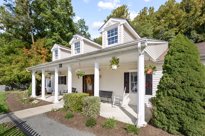 Front exterior view of a white house-style building with a covered porch supported by white columns. The porch has hanging flower pots, a wooden door with a wreath, a bench, and a rocking chair. The building is surrounded by green trees and shrubs under a partly cloudy sky.