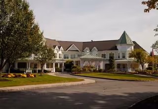 Front exterior of a two-story senior living building with turrets, wraparound porches, and a landscaped circular driveway.