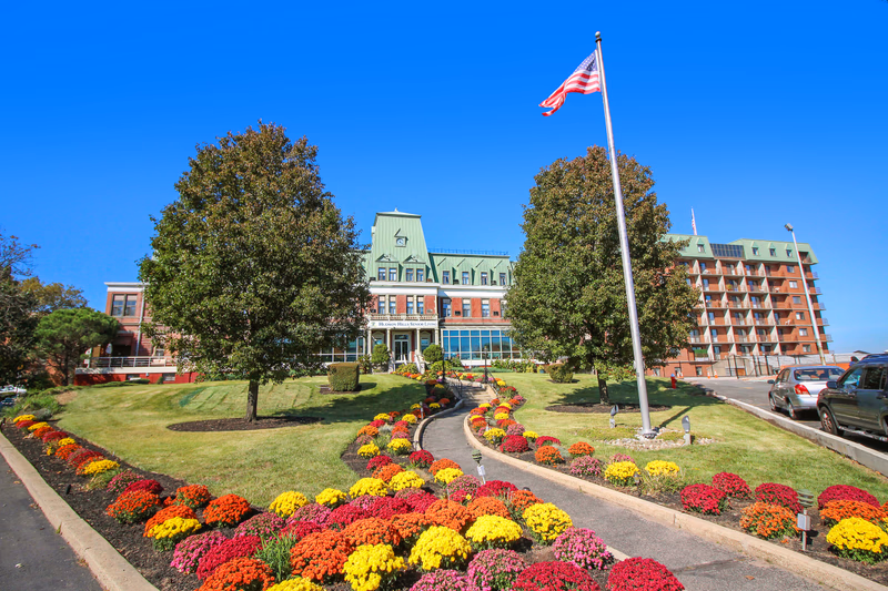 Front exterior view of Harbour View Senior Living Community building with a well-maintained garden featuring colorful flowers, two large trees, a flagpole with an American flag, and a clear blue sky.