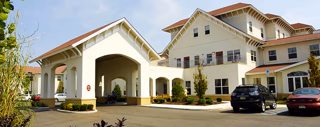 Front entrance of a multi-story senior living building with a covered porte-cochere, parked cars, and landscaped beds.