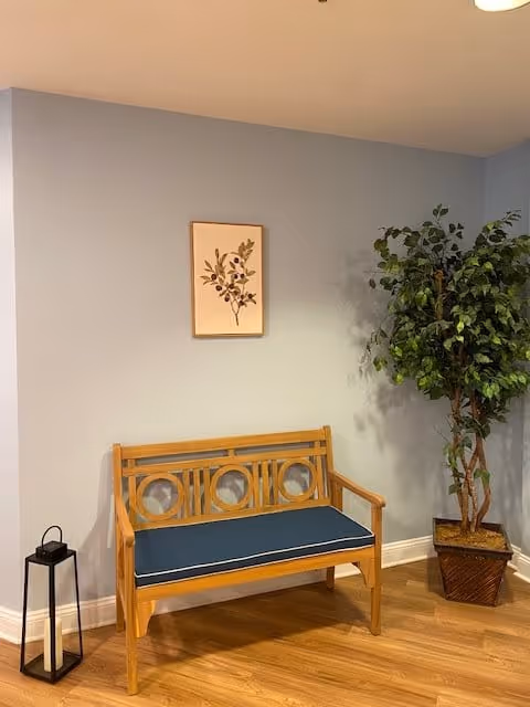 A small wooden bench with a blue cushion is placed against a light blue wall. Above the bench hangs a framed botanical print. To the right of the bench is a tall potted artificial plant with green leaves. On the floor to the left of the bench is a black lantern with a white candle inside. The floor is wooden.