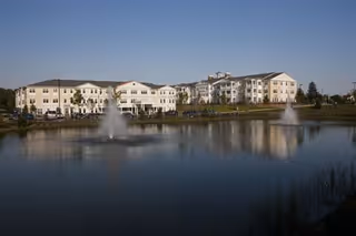 A large senior living facility building with multiple floors situated behind a pond with two water fountains, under a clear blue sky.