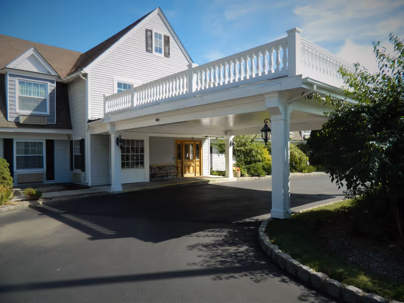 Exterior view of Colonial Manor At Panther Valley showing the entrance with a covered driveway supported by white pillars, a bench near the door, and a well-maintained garden area with shrubs and trees under a clear blue sky.