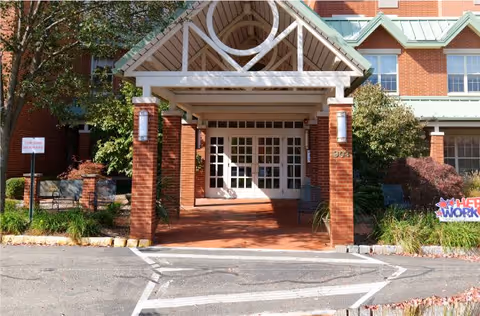 Entrance of a brick building with a covered porch supported by brick columns, white framed glass double doors, and surrounding greenery including bushes and trees.