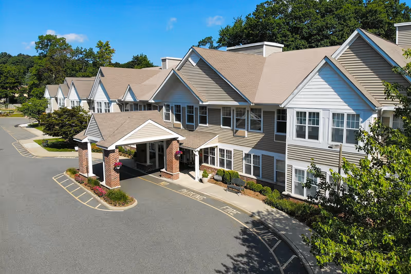 Exterior view of a senior living facility building with beige siding and white trim, featuring a covered entrance supported by brick columns. The driveway in front has marked fire lane and parking areas, with landscaped greenery and trees surrounding the building under a clear blue sky.