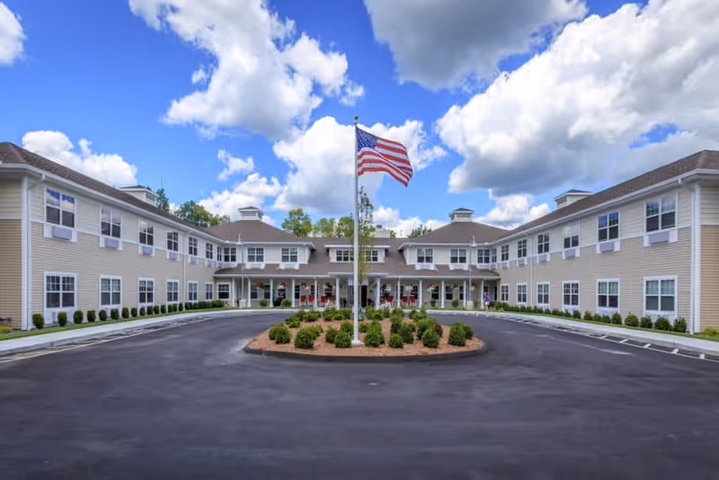 Front exterior view of a two-story assisted living facility building with beige siding and multiple windows. There is a circular driveway with a landscaped island in the center featuring small bushes and an American flag on a flagpole. The sky is blue with scattered clouds.