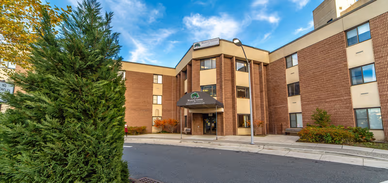 Exterior view of a multi-story brick building with a sign above the entrance that reads Majestic Center For Rehabilitation & Sub-Acute Care. The entrance has a covered awning and there is a street lamp nearby. Green bushes and a tree are visible in the foreground under a partly cloudy sky.