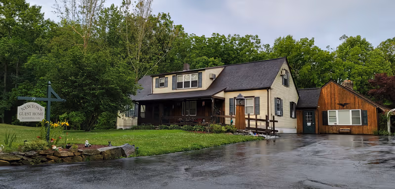 Two-story residential building with a covered front porch and adjacent wooden addition, a lawn sign reading 'Newton's Guest Home', and a wet driveway surrounded by trees.