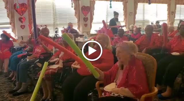 A group of elderly individuals seated in a well-lit room, participating in an activity using colorful foam sticks. The room is decorated with red heart-shaped decorations, suggesting a festive or themed event. A staff member is seen walking in the background.