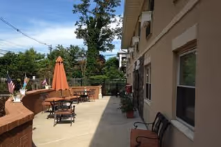 Outdoor patio area with several tables and chairs, some tables have umbrellas. The patio is adjacent to a building with windows and benches along the wall. Trees and greenery are visible in the background under a clear blue sky.
