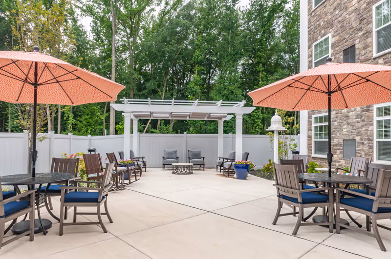 Outdoor patio with tables, orange umbrellas, chairs, a pergola, and potted plants beside a stone building.