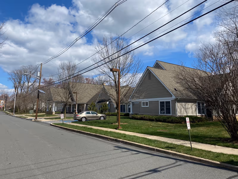 Street view of a senior living facility with beige buildings featuring pitched roofs, surrounded by leafless trees and green lawns under a partly cloudy blue sky. A silver car is parked near the buildings along the sidewalk.