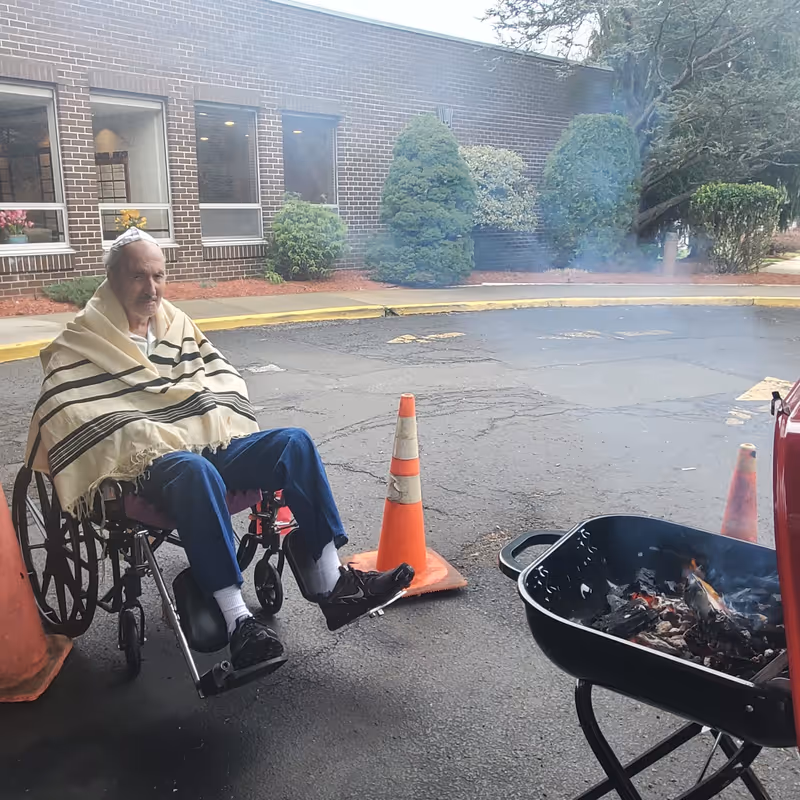 An elderly man in a wheelchair wrapped in a striped shawl sits outside near a brick building. There are orange traffic cones around him and a small grill with a fire burning inside it on the right side of the image.
