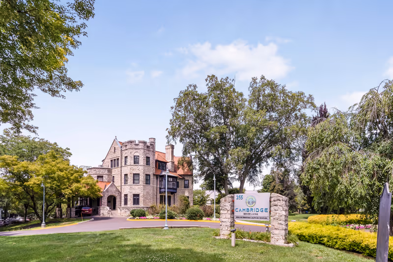 Exterior view of Cambridge Rehabilitation & Healthcare Center, a large stone building with castle-like architectural features surrounded by trees and greenery under a blue sky with some clouds. A sign in front displays the facility's name and address.